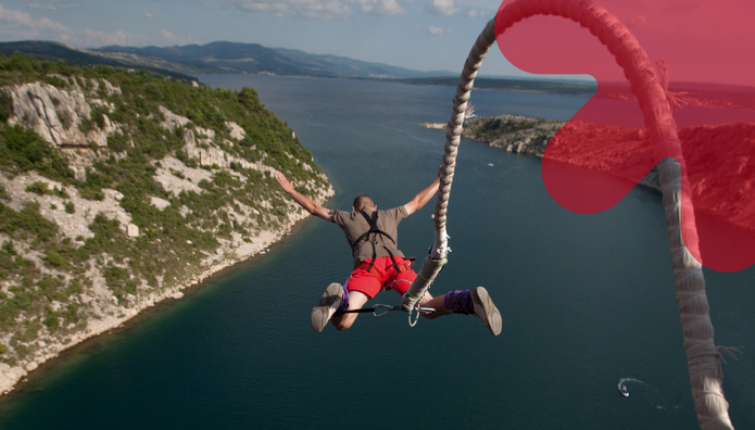 Mężczyzna w czerwonych szortach i uprzęży skacze na bungee z rozłożonymi szeroko rękami. Ujęcie od tyłu pokazuje skoczka zawieszonego nad rozległą zatoką otoczoną skalistymi, porośniętymi zielenią wzgórzami. Po prawej stronie widoczna jest gruba lina bungee, a w prawym górnym rogu – duży, półprzezroczysty czerwony symbol strzałki UniLodz.
