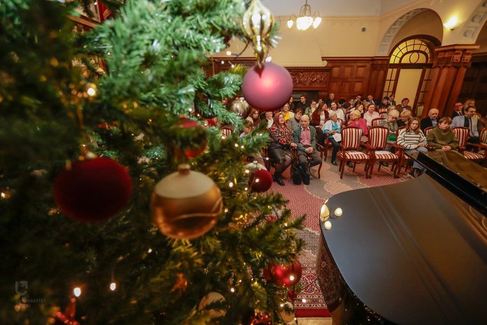 The photo shows the hall at the Biedermann’s Palace during a concert. A fragment of a Christmas tree, a piano, and a group of people sitting on chairs are visible.
