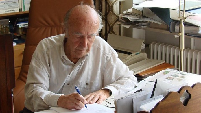 Karl Dedecius siedzi przy biurku z dużą liczbą papierów, książek i notatek, pisząc długopisem na kartce. Za nim znajduje się wysoki regał wypełniony książkami, a z boku wpada światło dzienne z okna. Scena przedstawia spokojną, skupioną pracę w otoczeniu pełnym materiałów naukowych.