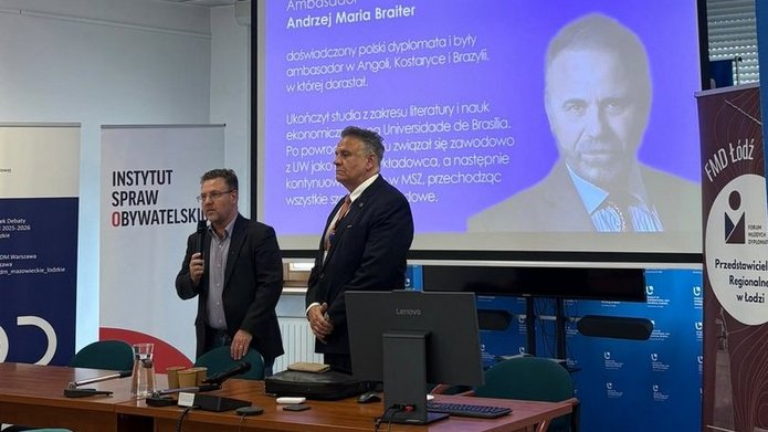 Osoba stojąca przy stole konferencyjnym przemawia do mikrofonu w sali spotkań, obok innej osoby stojącej przodem do zgromadzonych. W tle widoczny jest ekran z prezentacją zawierającą tekst: „Ambasador Andrzej Maria Braiter” oraz opis dotyczący polskiego dyplomaty i logo „Ministerstwo Spraw Zagranicznych”. Po lewej stronie ustawione są roll-upy, w tym „Instytut Spraw Obywatelskich”, a po prawej baner „FMD Łódź – Przedstawicielstwo Regionalne w Łodzi”. W pomieszczeniu znajdują się stoły konferencyjne, zielone krzesła, komputer Lenovo, mikrofony oraz naczynia z napojami. Nad salą widać sufit podwieszany z oświetleniem i zamontowany projektor.