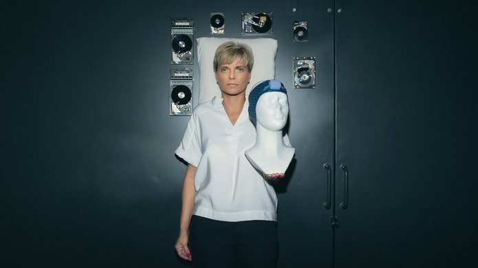 Prof. Renata Bocian holding a mannequin wearing a Vguard band, with a pillow and floating computer hard drives in the background